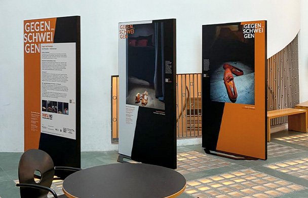 Drei Ausstellungstafeln mit dem Titel „Gegen das Schweigen“ in einem Foyer, Tisch im Vordergrund.